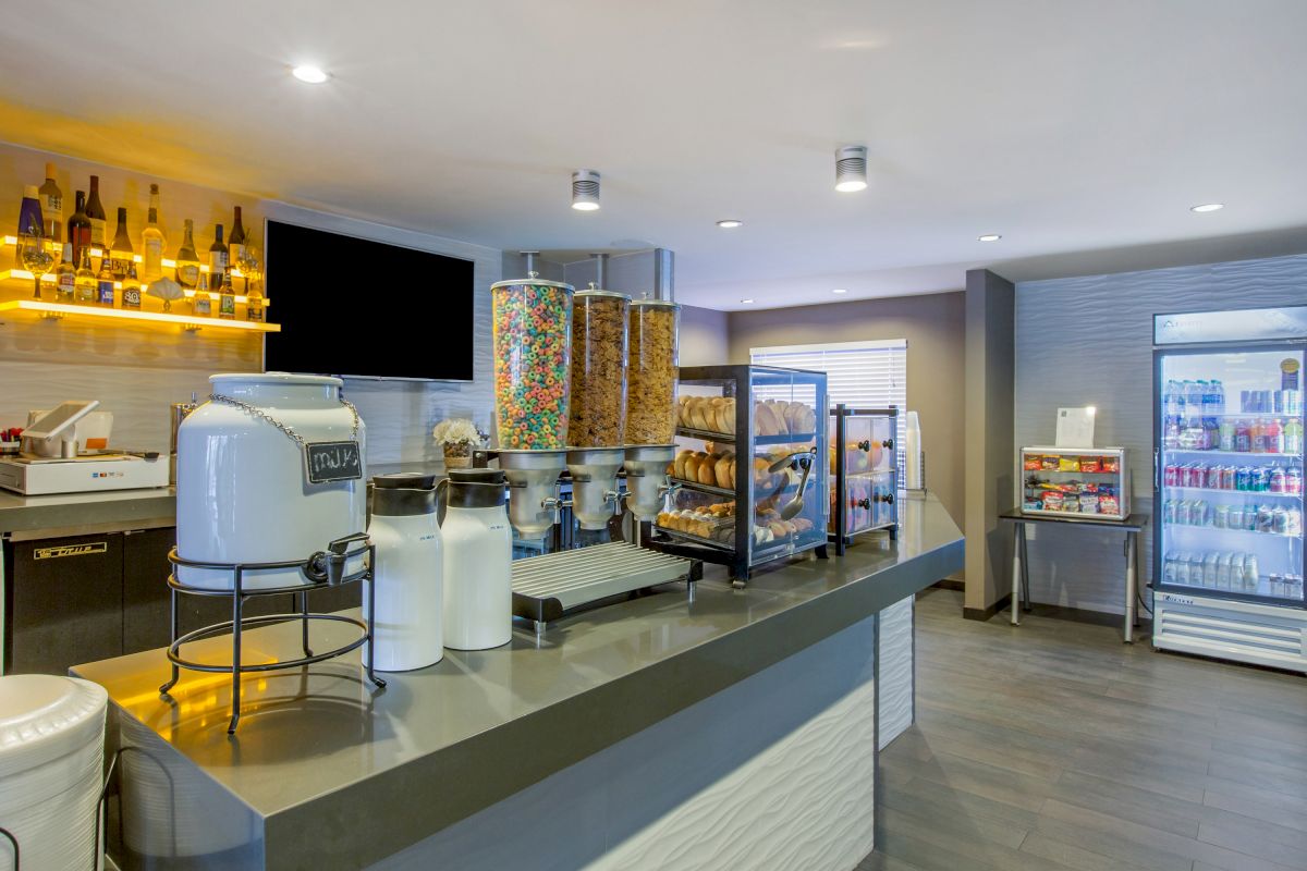 A self-service breakfast area with cereal dispensers, a beverage station, a fridge with drinks, and a selection of bread and bagels in trays.