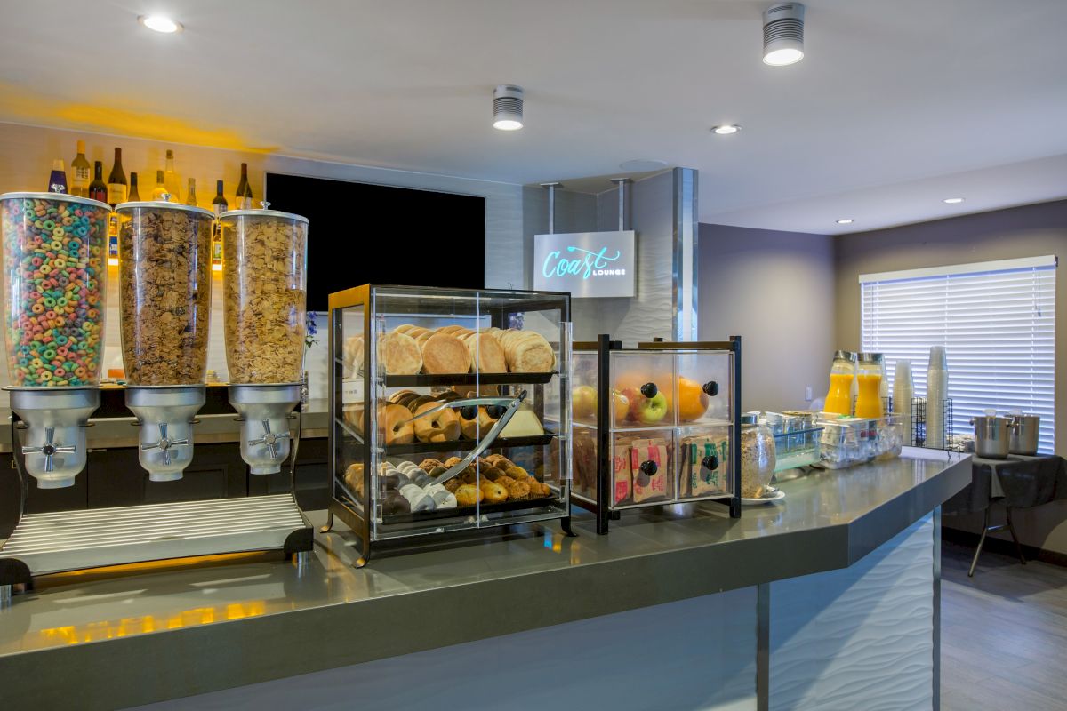 A breakfast buffet area with cereal dispensers, pastries, fruit, juice dispensers, and condiments on display.