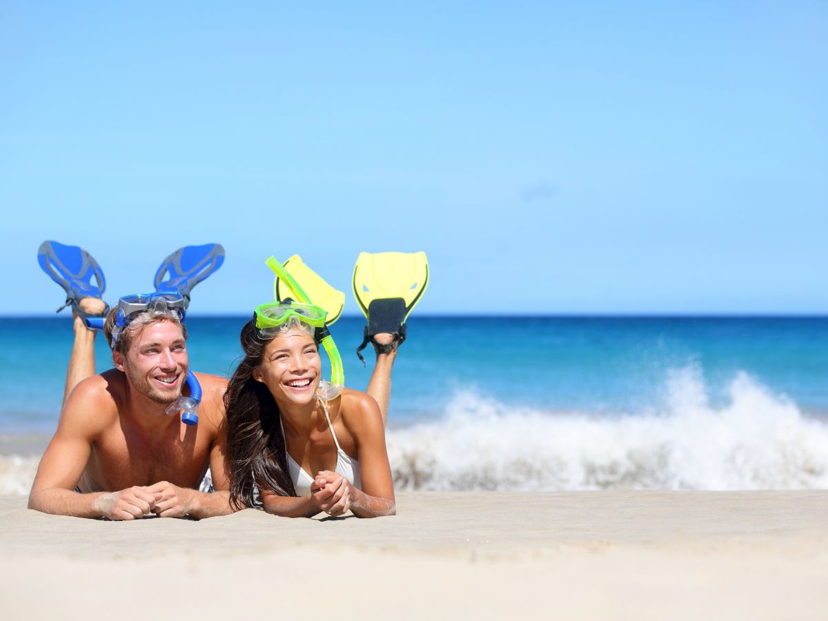 Two people in snorkeling gear lie on a sandy beach with the ocean and waves in the background, smiling and enjoying the sunny day.