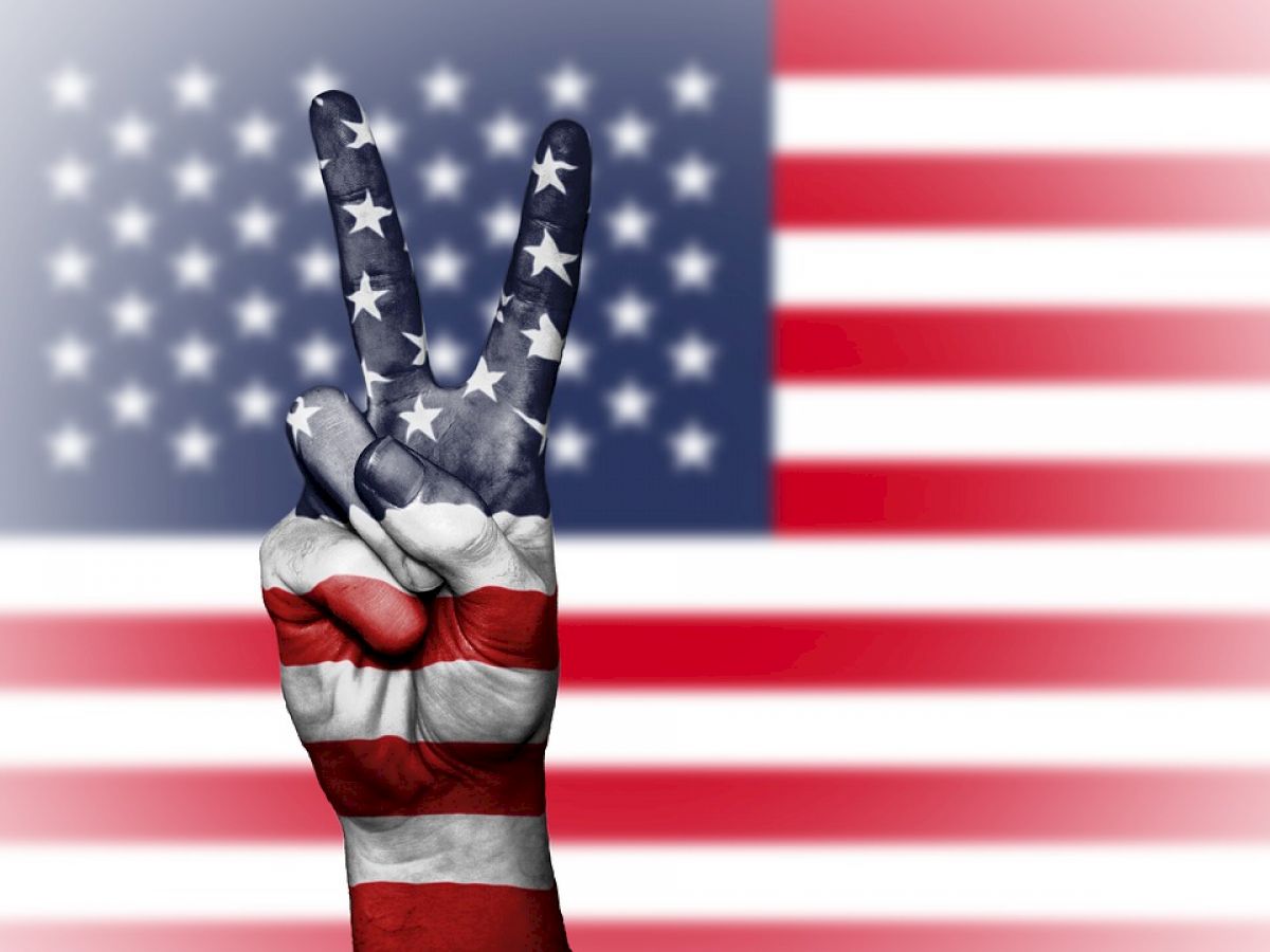 A hand painted with the American flag shows a peace sign in front of an American flag background.