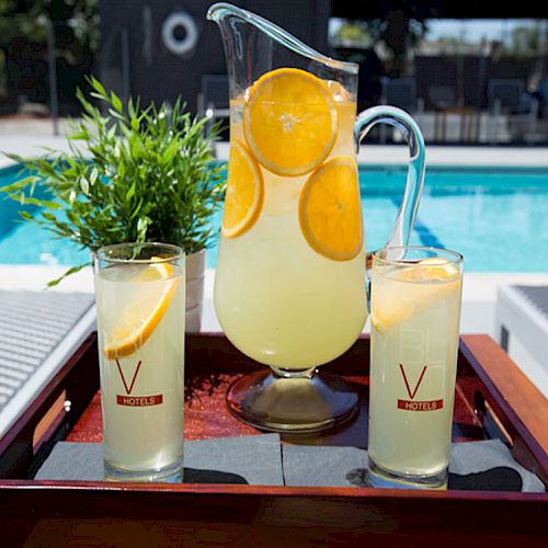 The image shows a pitcher of lemonade with orange slices and two glasses on a tray by a pool, accompanied by a potted plant.