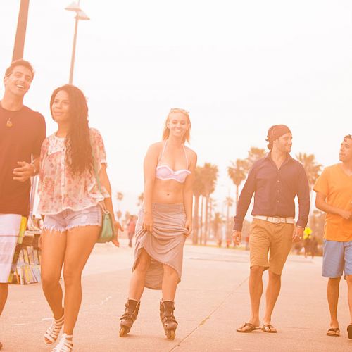 A group of people enjoying a walk on a sunlit pathway by the beach, with palm trees and buildings in the background.