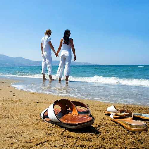 A couple holding hands walks along the beach shore, while two pairs of sandals are left on the sandy beach in the foreground.