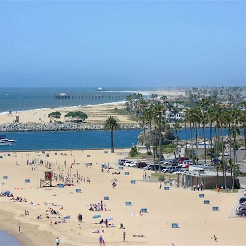 A beach scene with people sunbathing, umbrellas, parked cars, the ocean, palm trees, and a pier in the distance on a bright, sunny day.