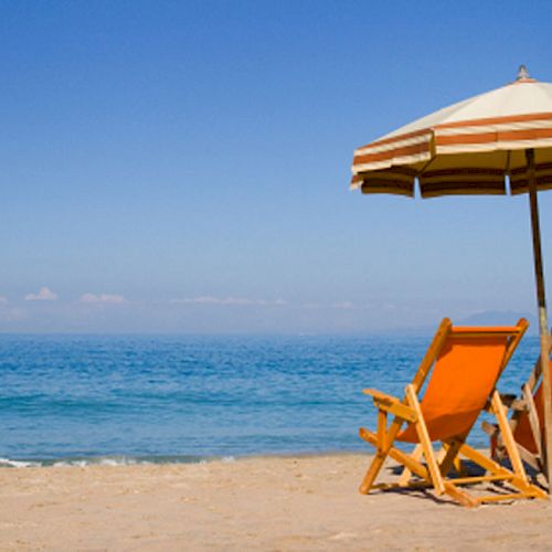 Two orange beach chairs sit under a striped umbrella on a sandy beach, facing the calm, blue ocean under a clear sky.