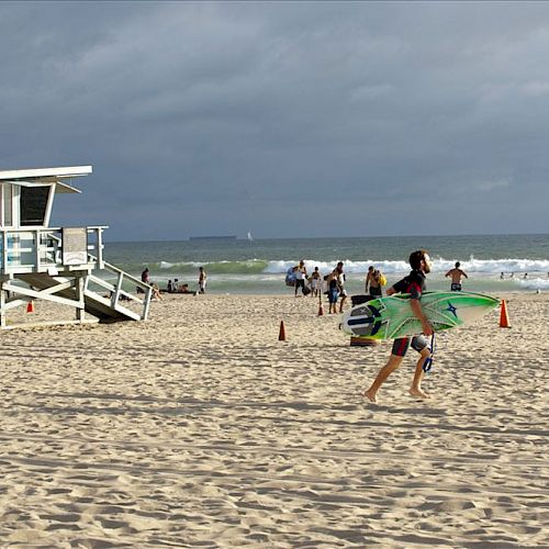 A beach scene with a lifeguard tower, people walking, and a person carrying a surfboard under a cloudy sky, ending the sentence.