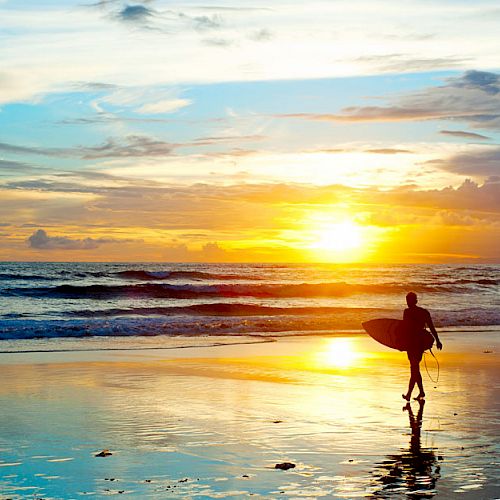 A surfer walks along the beach toward the ocean, carrying a surfboard, with a picturesque sunset casting vibrant colors over the water and sky.