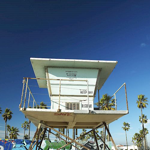 A lifeguard tower stands on a sandy beach, surrounded by palm trees, against a clear blue sky.