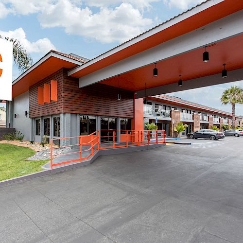 The image shows the exterior of a hotel with a parking area and signage reading 