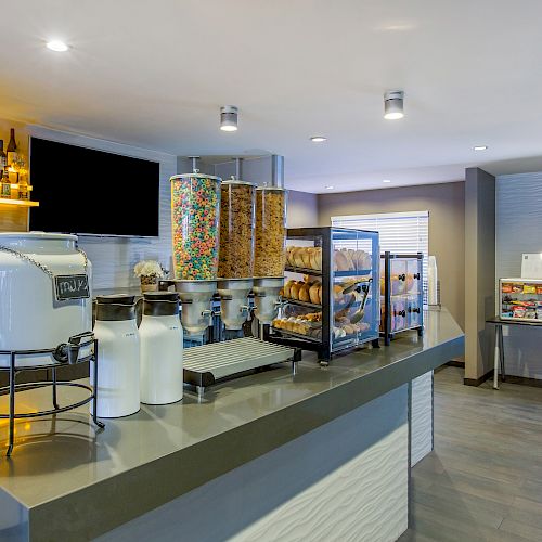 A breakfast buffet area featuring cereal dispensers, a bagel display, milk dispensers, a mini fridge with drinks, and assorted snacks on shelves.