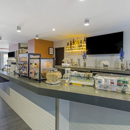 The image shows a modern breakfast bar with a variety of food and drinks, including cereal and juice, and seating areas in the background, well-lit.