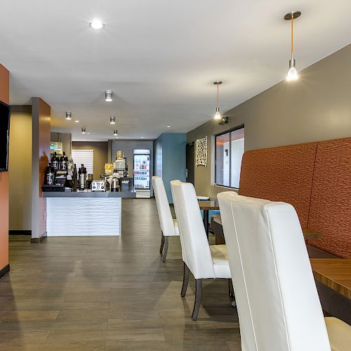 A modern dining area with white chairs, a wall-mounted TV, hanging lights, and a counter in the background for serving food and beverages.