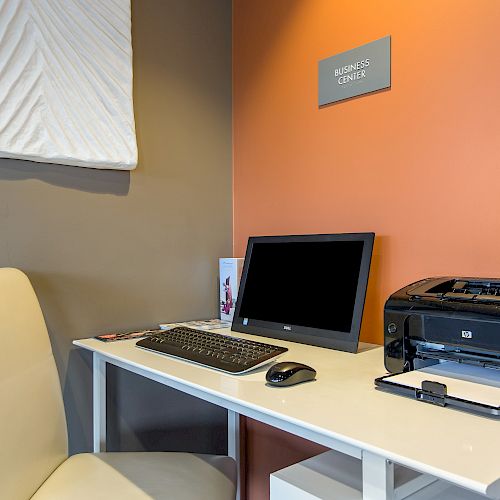 A small office setup with a computer, keyboard, mouse, and printer on a white desk, next to a white chair and a sign that reads 