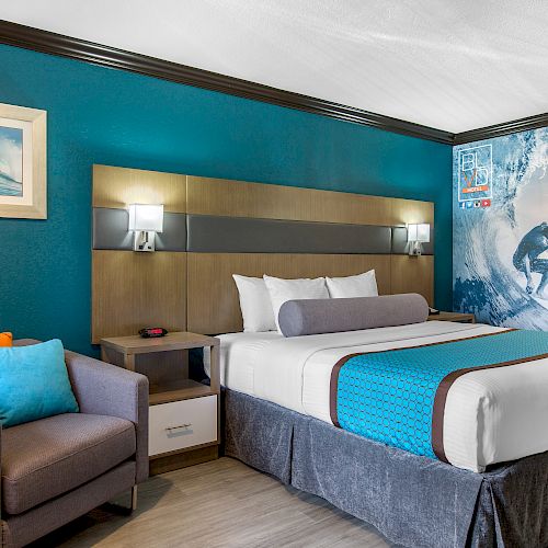 A modern hotel room features a bed with teal accents, a gray couch with orange and teal cushions, and surfing-themed wall art, creating a beach vibe.