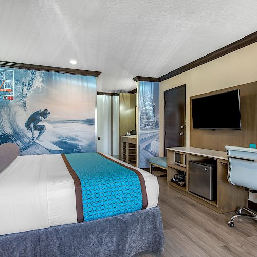 A modern hotel room with a surfing theme, featuring a bed, desk, chair, TV, mini-fridge, and surf-themed wall art.