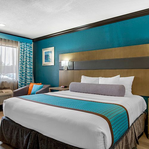 A hotel room with a large bed, teal walls, a window, nightstands, and modern decor, including lamps and a wall frame.