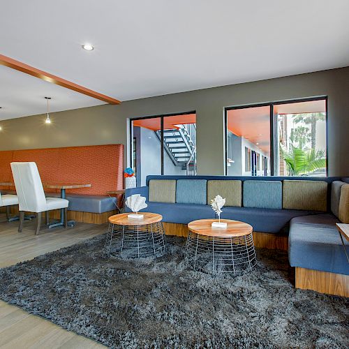 This image shows a modern lounge area with a gray rug, comfortable seating, tables, decorative flowers, and large windows letting in natural light.