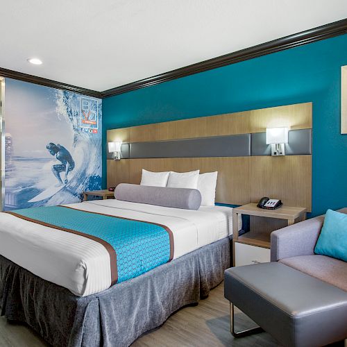 The image shows a modern hotel room with a surfer-themed decor, including a bed, couch, and wall art depicting a surfer on a wave.