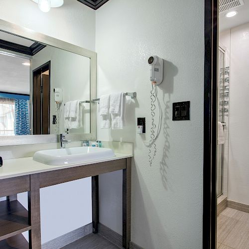 A hotel bathroom with a large mirror, a sink, towels, a hairdryer, and a toilet. Toiletries are visible, and there's a window and blue curtains.