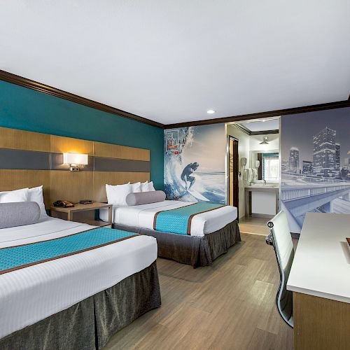 A hotel room features two double beds with teal accents, a mural on the wall, a flat-screen TV, a desk with a lamp, and bathroom access.