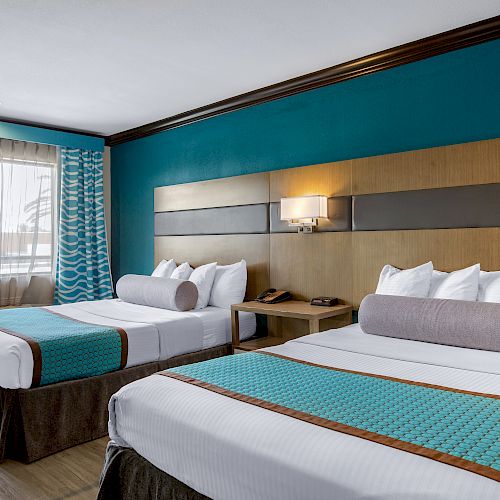 A hotel room with two neatly made double beds, teal accents, a window with drapes, wall lighting, and a bedside table with a phone and lamp.