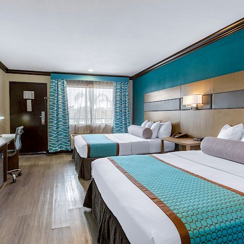 This image shows a hotel room with two large beds, a flat-screen TV, a desk, and blue accents on the bedding and curtains.