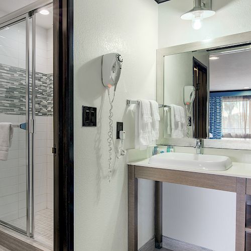 A bathroom with a glass shower, sink, mirror, hairdryer, and towels. Beyond, a room with a bed and blue curtains is visible.