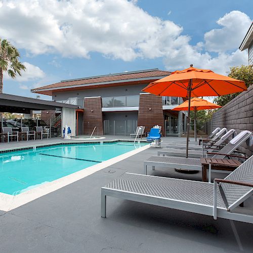 The image shows an outdoor swimming pool with lounge chairs, orange umbrellas, and a modern building in the background under a partly cloudy sky.