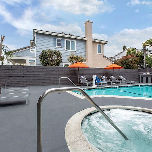 The image shows an outdoor swimming pool and hot tub, surrounded by lounge chairs and orange umbrellas, with houses and blue sky in the background.