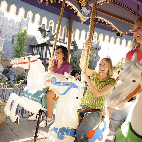 Three people are enjoying a ride on a carousel with white horses, near a castle-like structure, smiling and having fun in the sun.