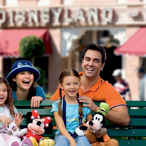 A family of four sits on a bench at Disneyland holding Mickey Mouse and Goofy plush toys, with the park's entrance visible in the background.