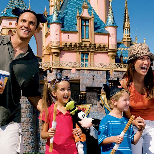 A family of four is happily posing in front of a castle at a theme park, holding snacks and drinks, wearing themed hats.