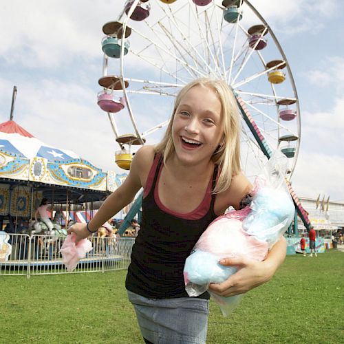 A girl is smiling and holding cotton candy at an amusement park with a Ferris wheel and a carousel in the background.