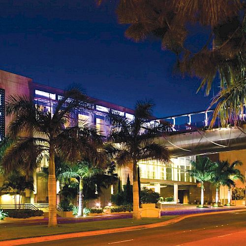 The image shows a night view of the South Coast Plaza shopping center with palm trees and a pedestrian bridge, lit up with lights.