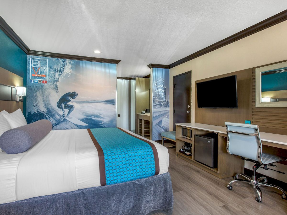 A modern hotel room with a surf theme, featuring a bed, desk, chair, TV, and refrigerator, with surf-themed wall art and blue accents.