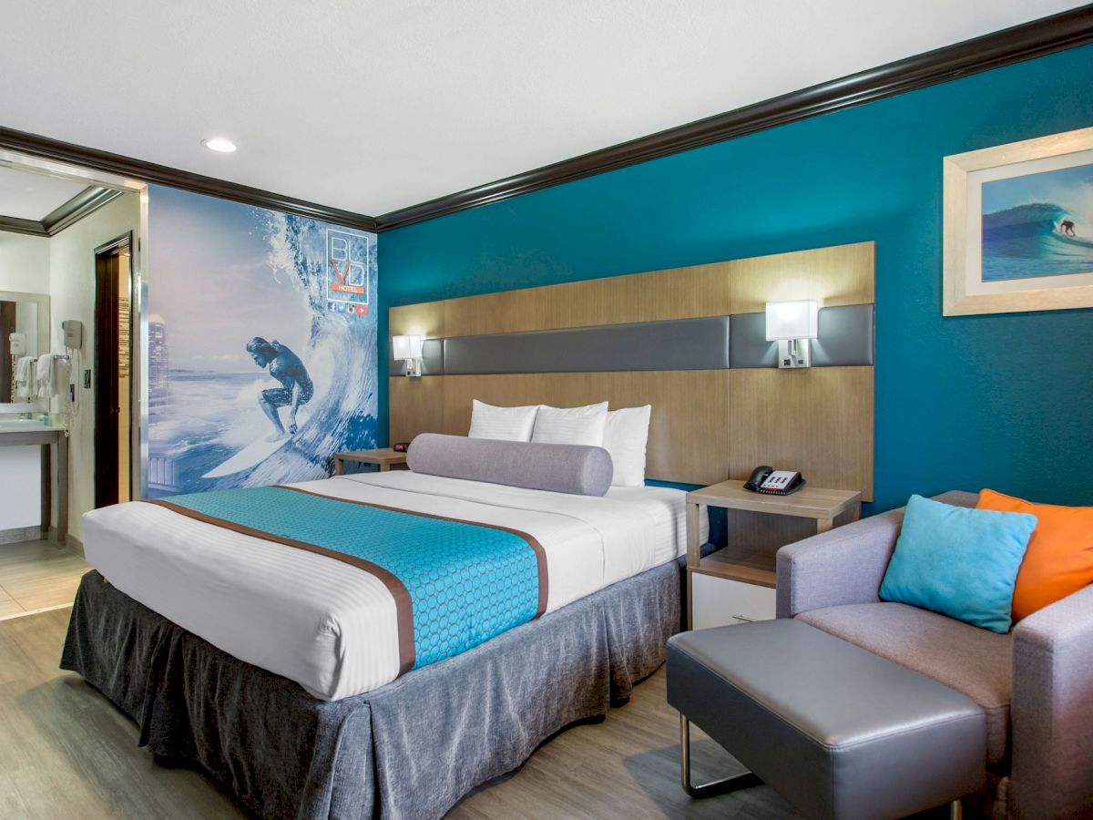 A modern hotel room with a surfing theme, featuring a bed, armchair, wall art, bedside table, and a view of the bathroom entrance.