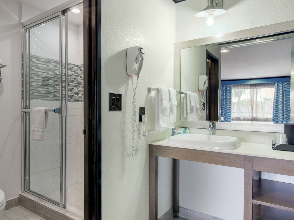 The image shows a modern bathroom with a walk-in shower, a wall-mounted hairdryer, a sink with a large mirror, and a glimpse of an adjoining room.