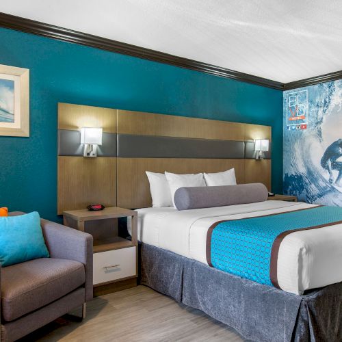 A modern hotel room with a surf theme, featuring a bed, a nightstand, a wall with a surfing mural, a framed surf photo, and a cozy chair.