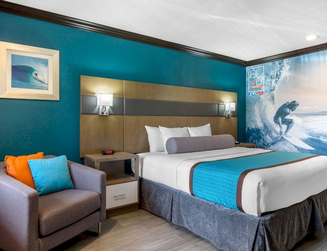 A modern hotel room with a surf theme, featuring a bed, a nightstand, a wall with a surfing mural, a framed surf photo, and a cozy chair.