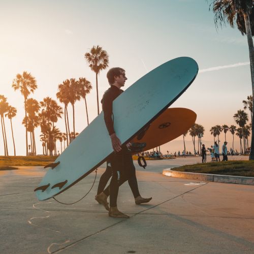 Two surfers in wetsuits carry their surfboards along a palm tree-lined path, possibly heading to the beach for a surf session at sunset.