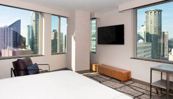 The image showcases a modern hotel room with large windows offering a cityscape view, a bed, a chair, a TV, and minimalist furnishings.