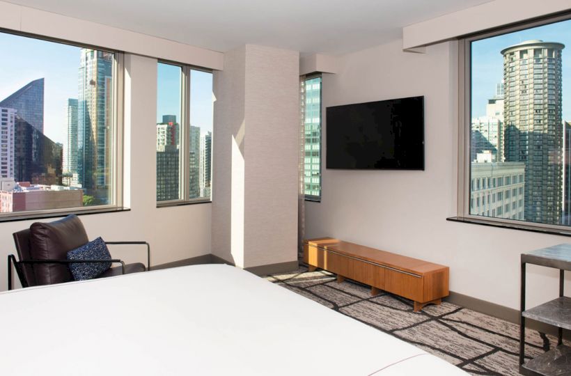 The image showcases a modern hotel room with large windows offering a cityscape view, a bed, a chair, a TV, and minimalist furnishings.