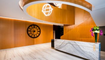 The image shows a modern lobby with a marble reception desk, a large clock on a wooden wall, and contemporary lighting fixtures.