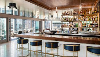 A modern bar with a long wooden counter, elegant barstools, hanging lights, and a well-stocked back wall of various alcohol bottles and glassware.