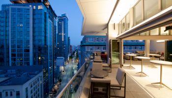 The image shows an outdoor balcony with chairs and tables, overlooking a cityscape with tall buildings and a street below, captured at twilight.