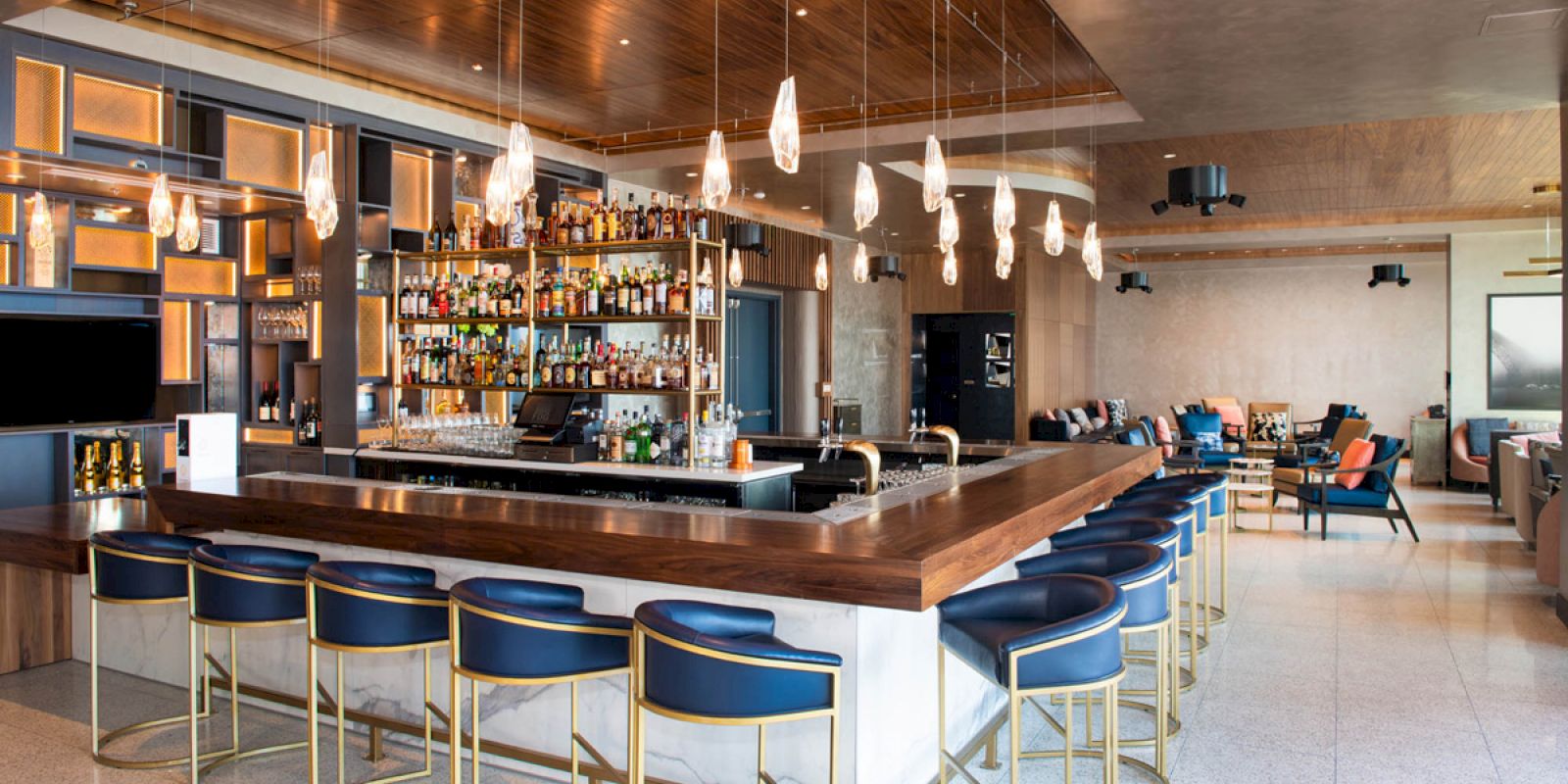 This image shows a modern bar with a wooden countertop, numerous liquors on shelves, blue padded bar stools, stylish lighting, and a seating area.