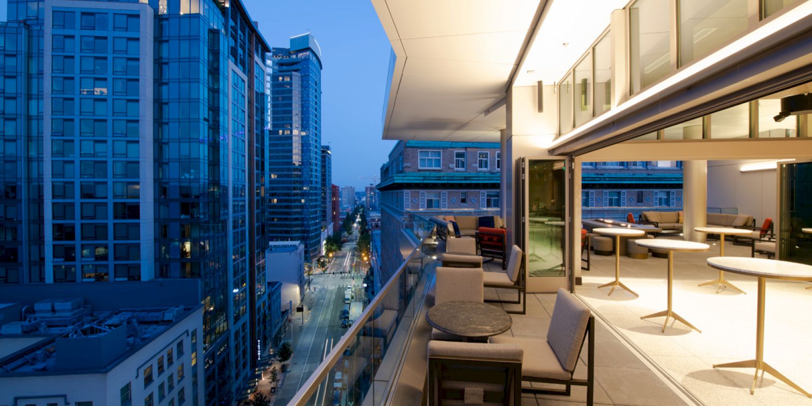 A modern cityscape at twilight, featuring tall buildings and a sleek balcony with tables and chairs overlooking a busy street.