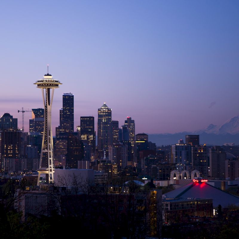 City skyline at dusk featuring the Space Needle, skyscrapers, and Mount Rainier in the distance.