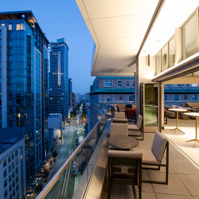 A modern urban balcony at twilight with comfortable seating and tables, overlooking a street with tall buildings and city lights.