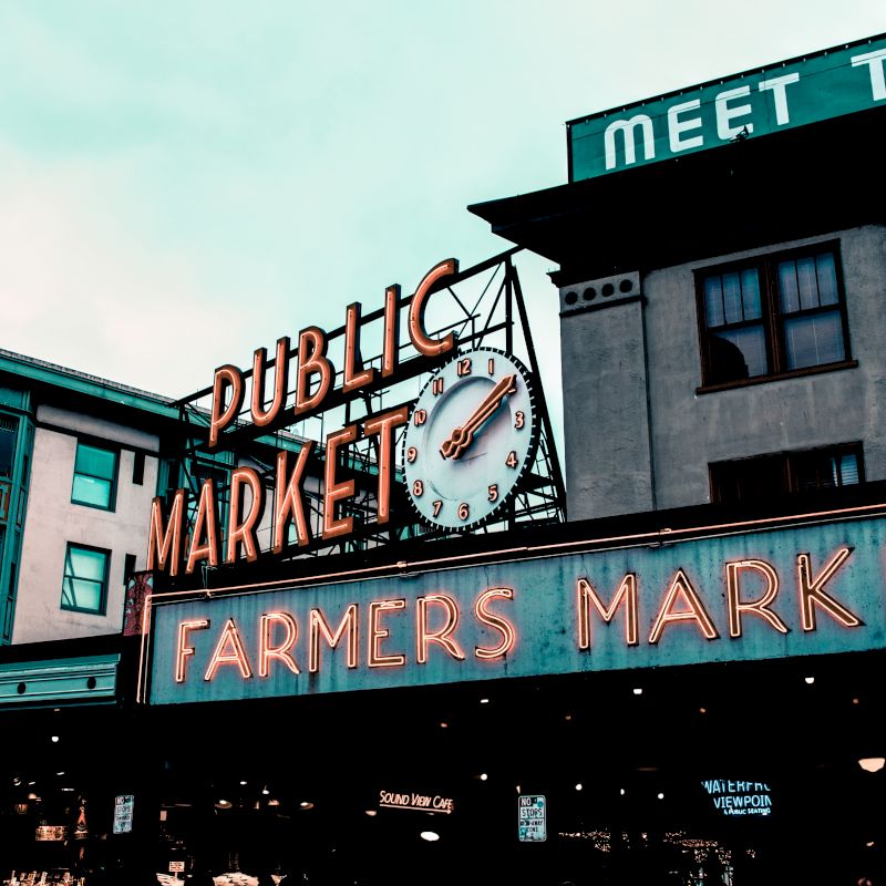 The image shows the iconic neon sign of a "Public Market" and "Farmers Market" with a clock. The area has historic buildings and bustling activity.